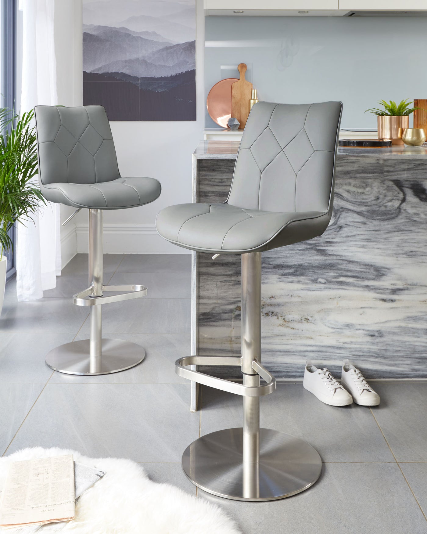 Two modern grey bar stools with quilted leather upholstery and stainless steel pedestal bases with integrated footrests.