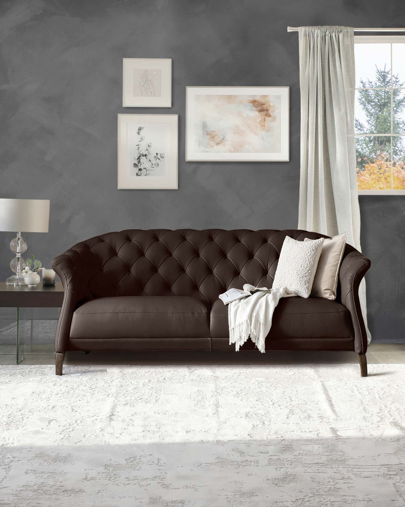Elegant chocolate brown leather upholstered sofa with a classic tufted backrest and sleek curved armrests. Accompanied by soft white accent pillows and a fringe throw blanket, resting on a distressed white area rug. A glass-top side table with a modern table lamp is positioned to the left.