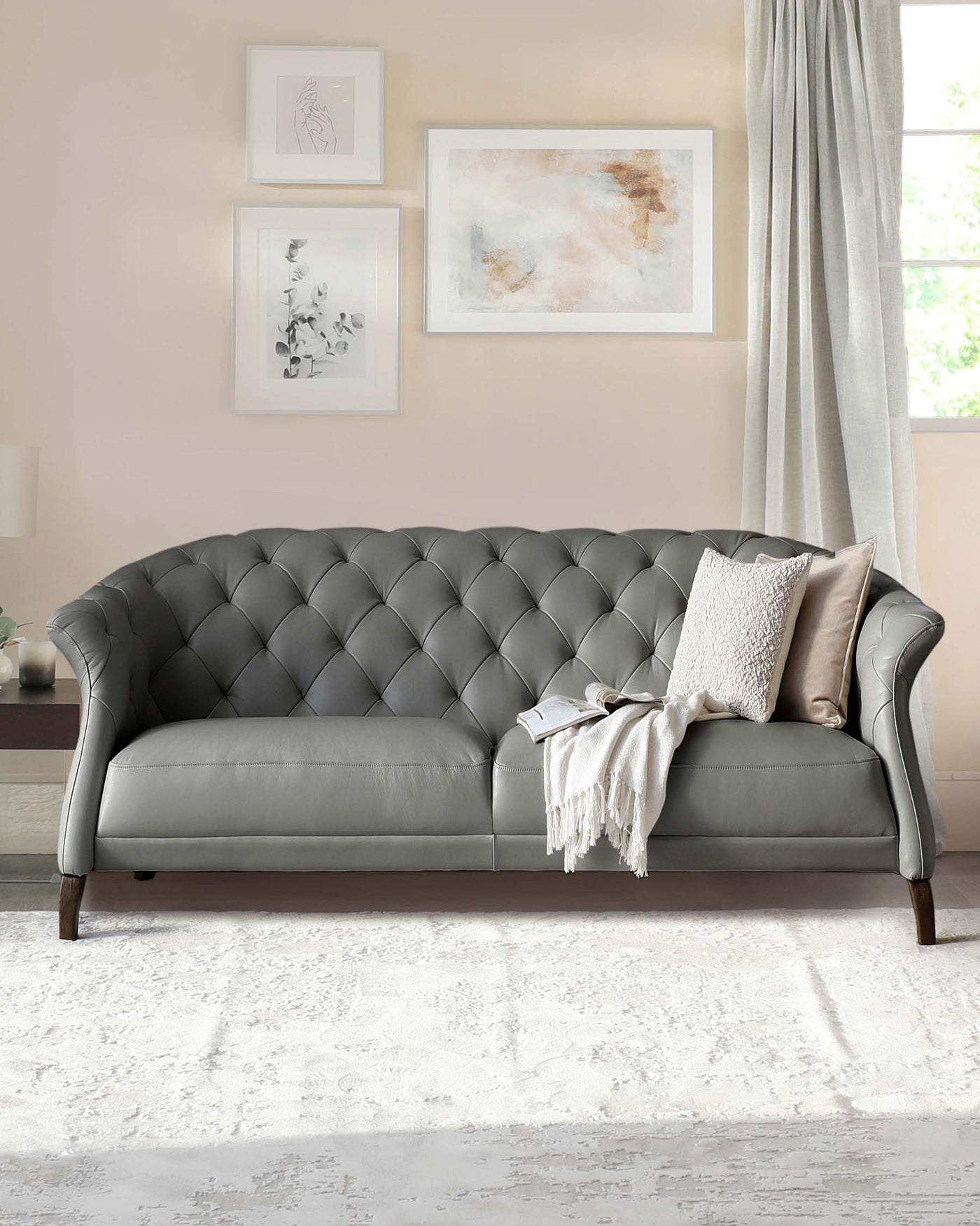 Elegant grey tufted Chesterfield sofa with rolled arms and wooden legs, adorned with two throw pillows and a cream blanket, set against a neutral-toned wall with framed artwork, above a textured white area rug.