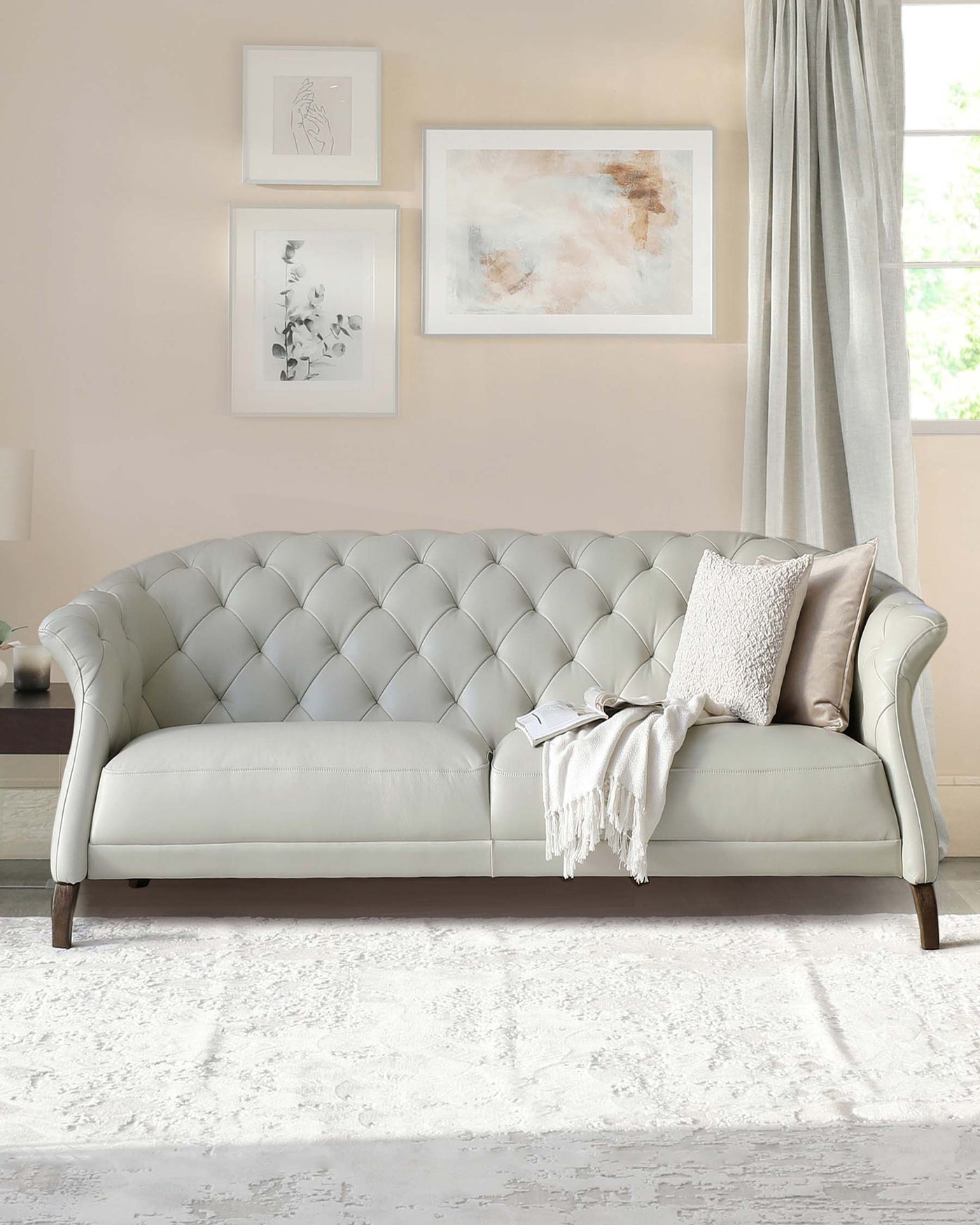 Elegant, tufted, light beige fabric sofa with scrolled arms and dark wooden legs, adorned with a pale pink pillow and a white fringed throw blanket, set in a softly lit room with a cream area rug.