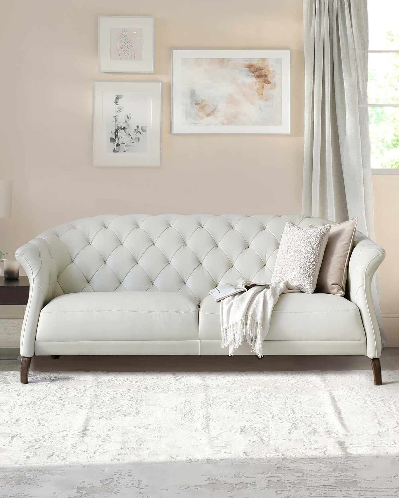 Elegant ivory tufted chesterfield sofa with rolled arms and dark wooden legs, adorned with a textured throw blanket and decorative pillows, set against a light patterned area rug in a room with neutral wall colours and framed artwork.