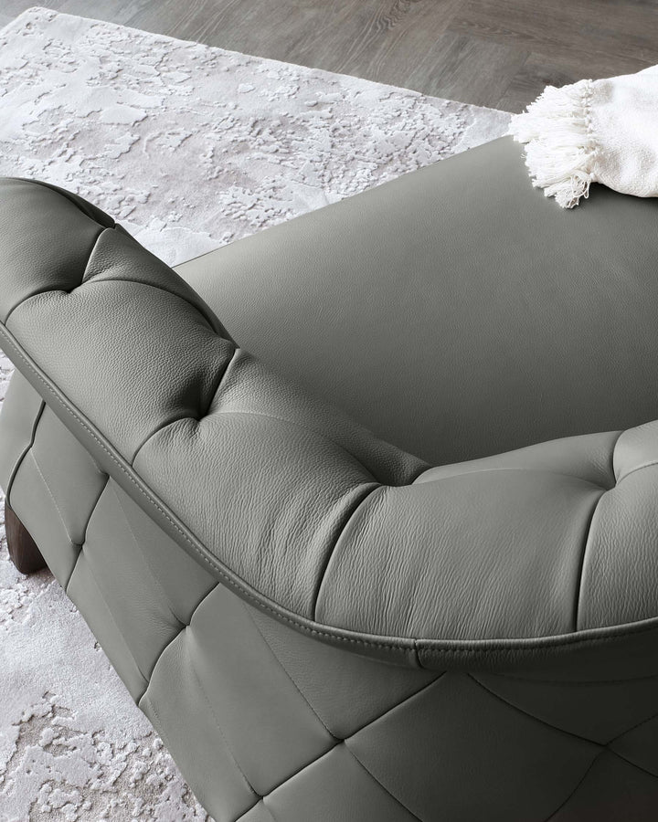 Modern charcoal grey leather sofa with tufted backrest and cushioned armrests, featuring elegant stitching details, placed on a textured white and grey area rug, in a room with wooden flooring.