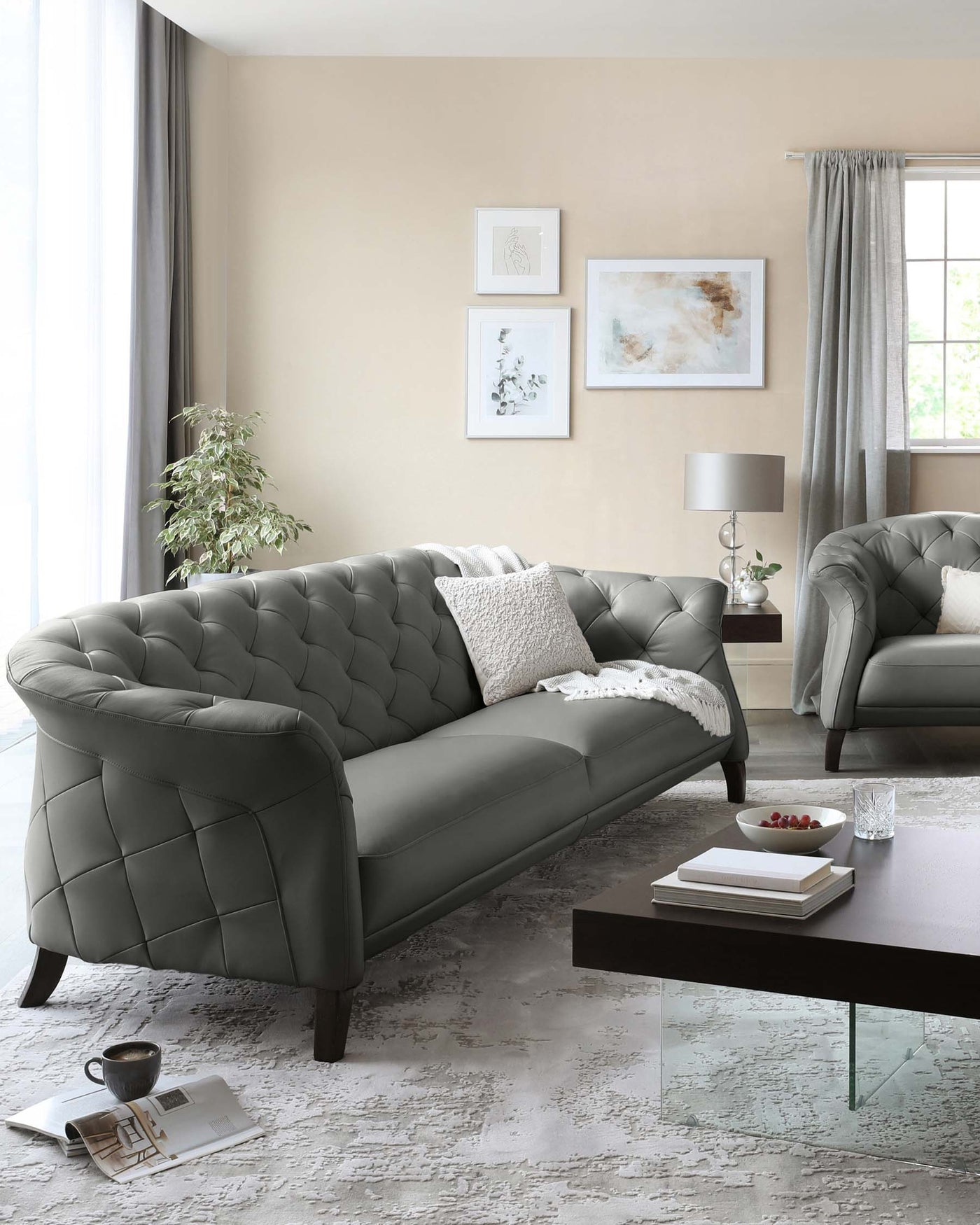 Elegant living room set featuring a tufted dark grey sofa with matching armchair, complemented by a modern dark brown rectangular coffee table with a glass bottom shelf. The room is accented with a soft textured area rug, simplistic white-and-silver decorative pillows, and a small round side table supporting a contemporary lamp with a grey shade.