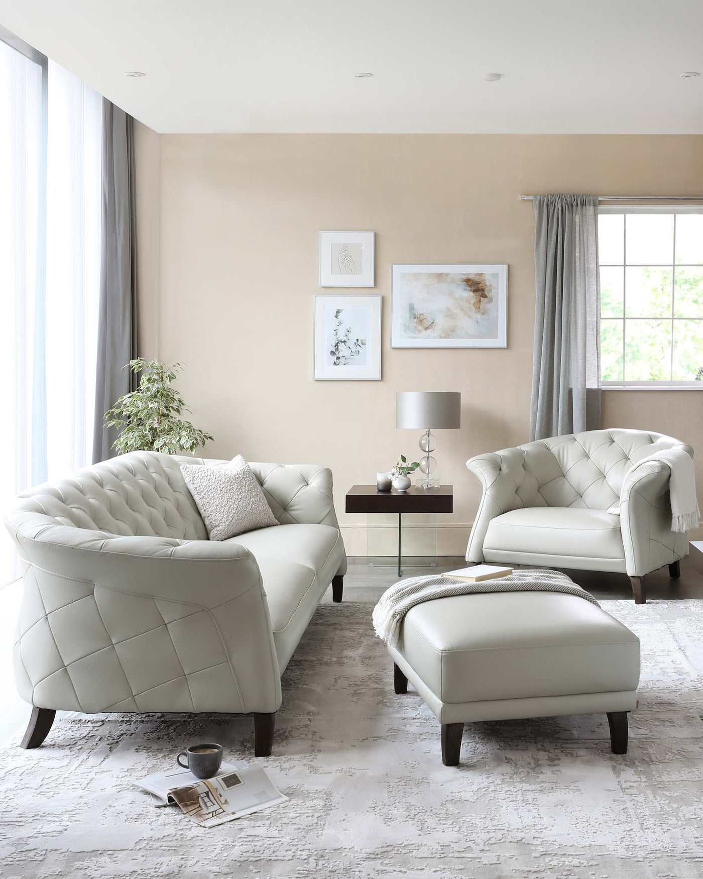 Elegant living room ensemble featuring a tufted cream-colored sofa with matching armchair, both with a quilted diamond-pattern backrest and curved arms. A coordinating ottoman rests in front, atop a textured white and grey area rug. A small rectangular wooden side table with a modern lamp sits beside the armchair. The room is accented with minimalist wall art, grey drapery, and a potted indoor plant.
