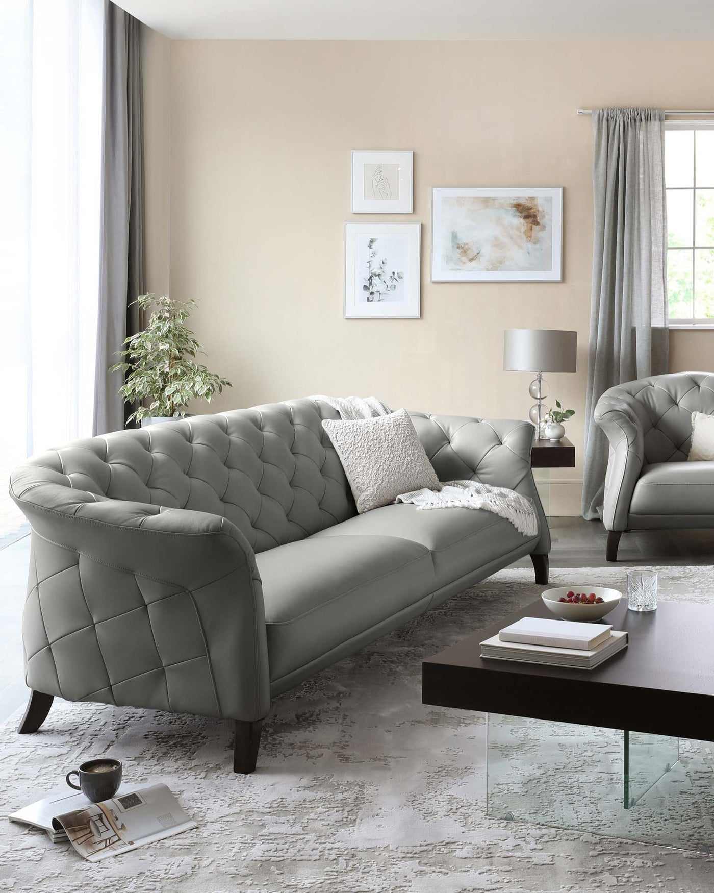 Elegant, grey tufted chesterfield sofa with matching armchair, featuring a plush design with deep button accents. In front, a modern, low-profile, rectangular coffee table with a dark wood finish and glass legs. The room enhances the sophisticated look with a textured white area rug, and a side table holding a lamp with a grey shade.