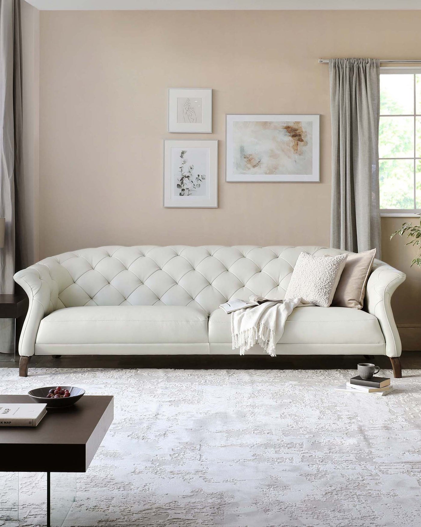 Elegant ivory tufted chesterfield sofa with rolled arms and wooden legs, complemented by a textured beige throw and decorative cushion. In the foreground, a modern square side table with a dark finish displaying a bowl of fruit and a hardcover book.