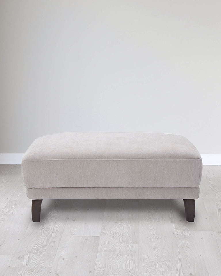 Modern minimalist light grey fabric upholstered ottoman with sturdy dark wooden legs on a light hardwood floor against a plain white wall.