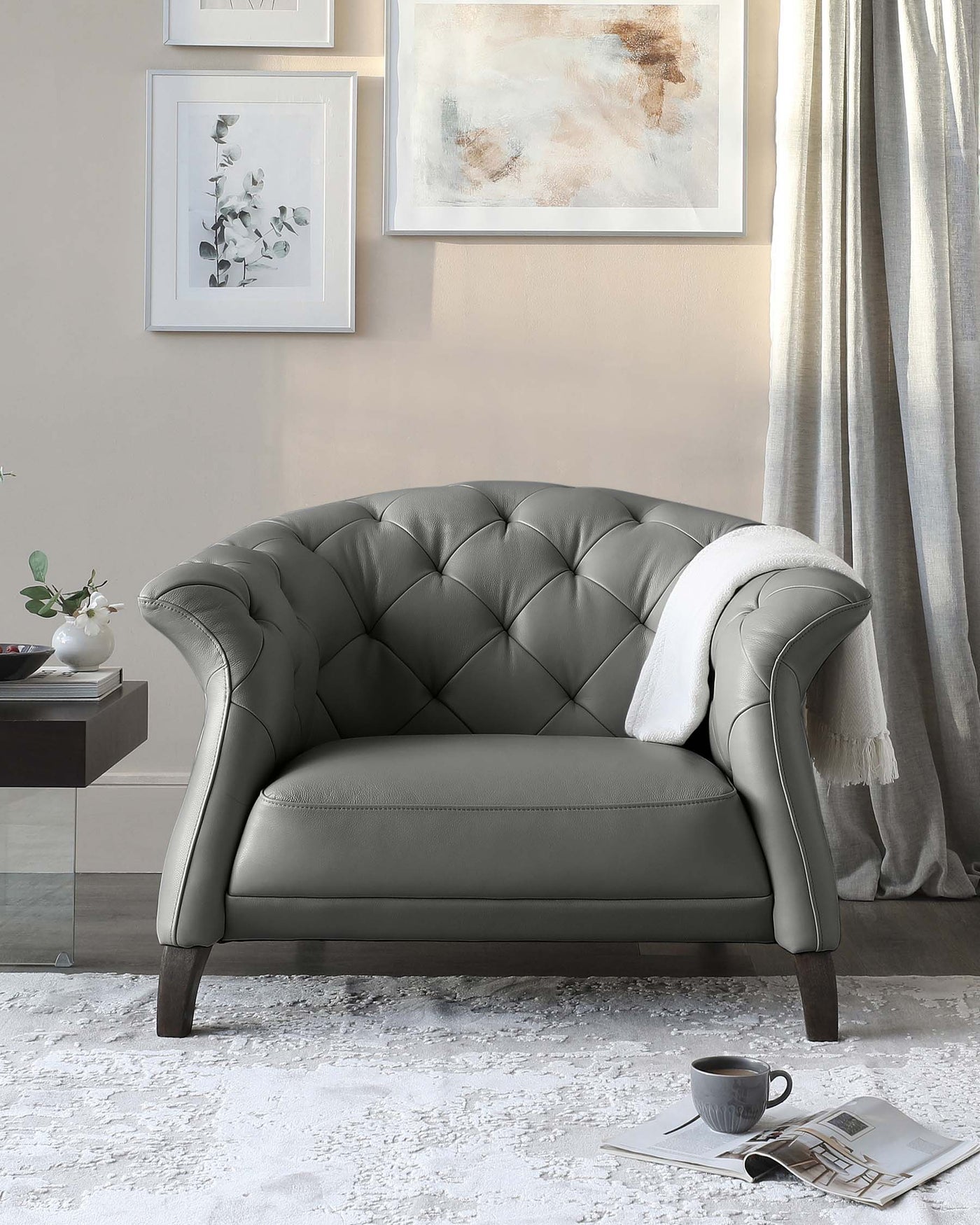 Elegant grey tufted loveseat with a classic chesterfield backrest, featuring dark wooden legs, accented with a white throw blanket draped over one arm, set against a neutral-toned living room backdrop with framed artwork, a silver-grey curtain, and a plush white area rug, with a coffee cup and magazine resting on the rug.
