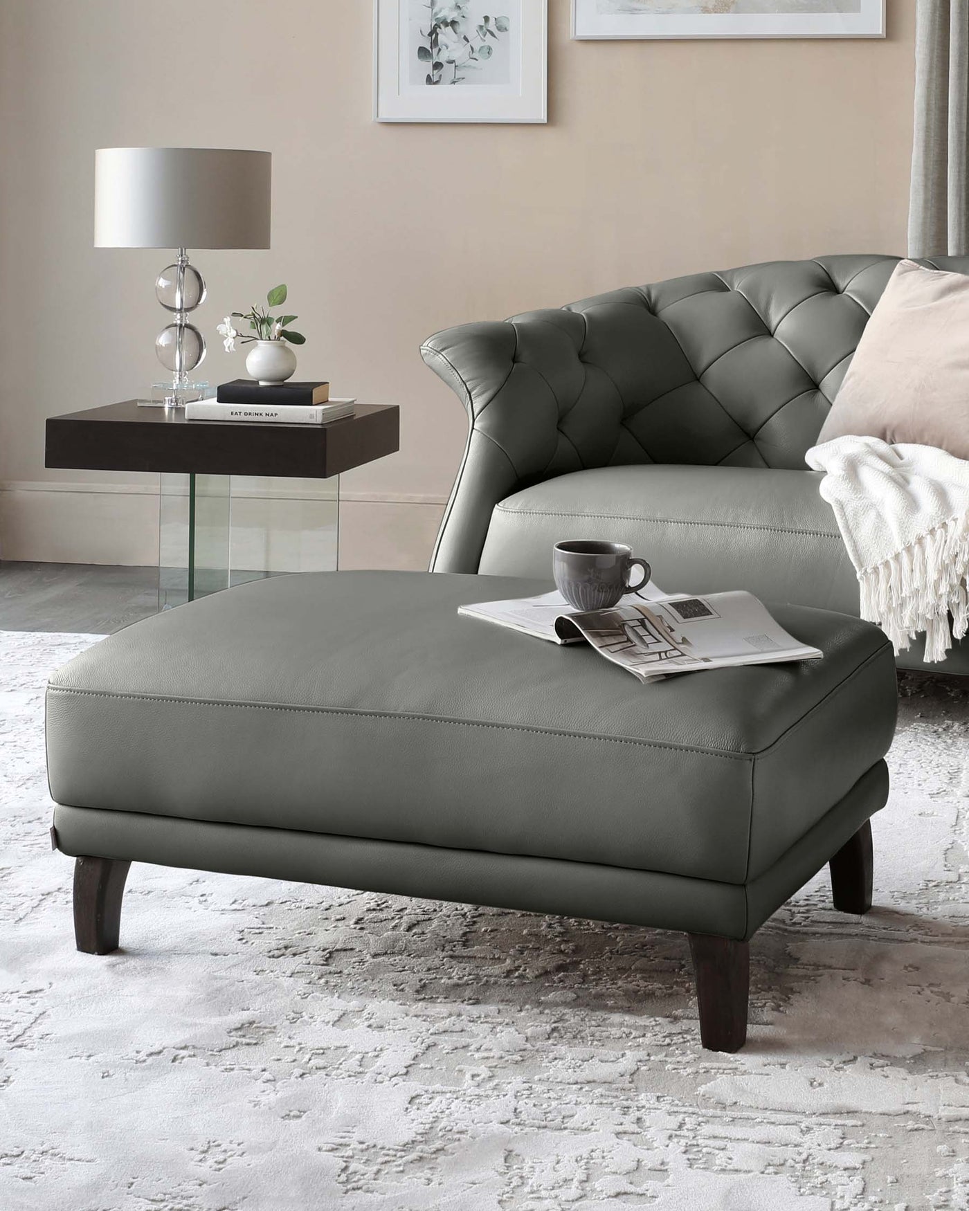 A luxurious grey leather tufted Chesterfield loveseat with a matching oversized ottoman, complemented by a modern glass and dark wood side table displaying decorative items and a contemporary style lamp.