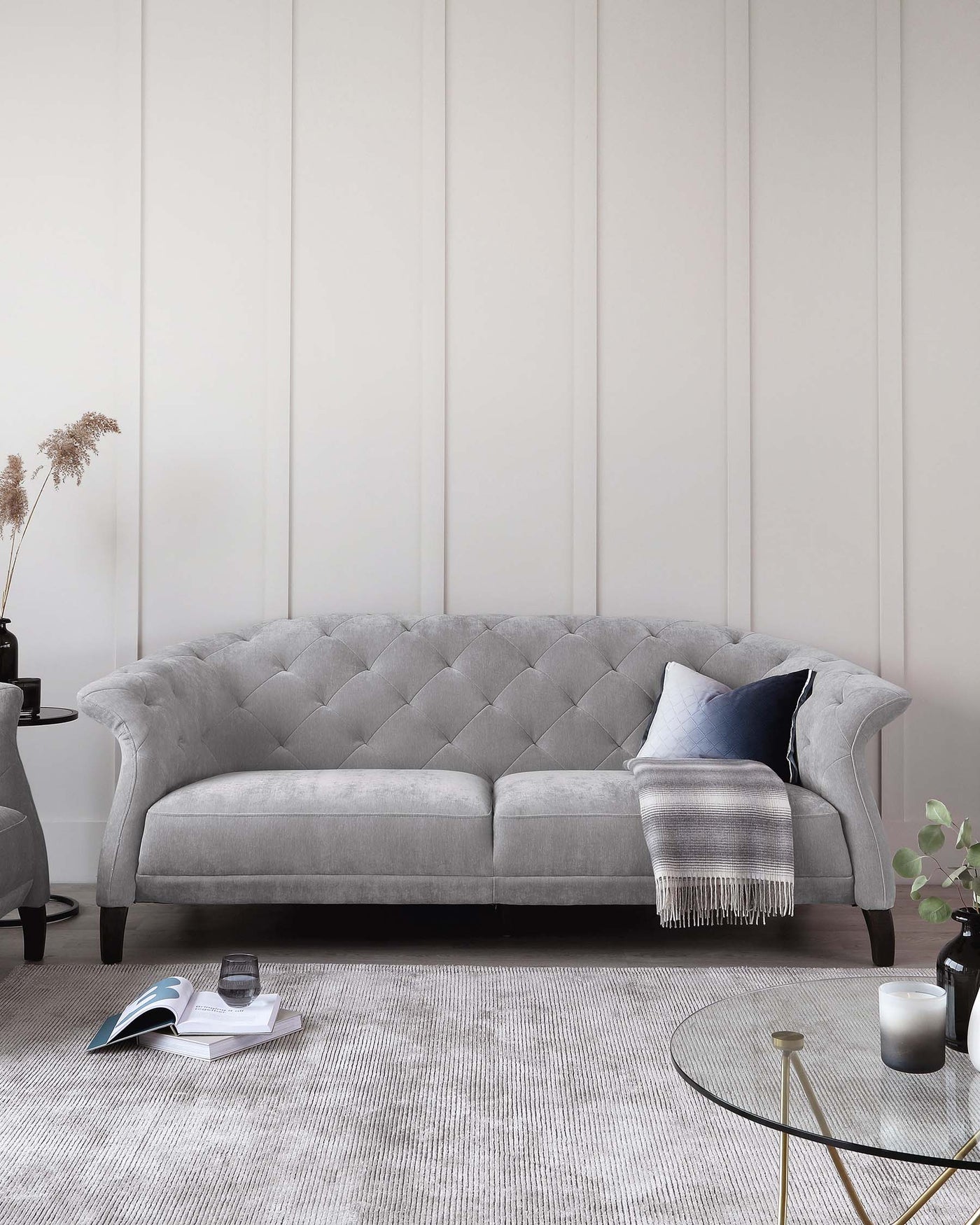 Elegant light grey tufted chesterfield sofa with accent cushions and a fringed throw blanket, flanked by round black side tables with vases and books, set on a textured white area rug, next to a glass-top coffee table with gold accents.