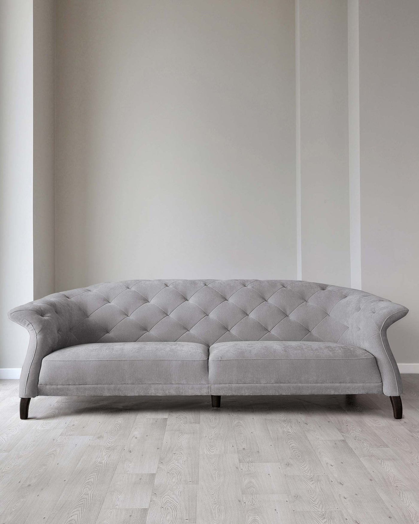 Elegant grey tufted fabric sofa with a curved silhouette and low backrest, standing on wooden legs against a neutral wall on a light hardwood floor.