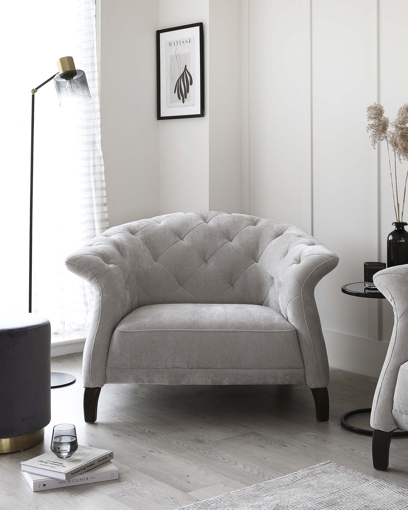 Elegant grey tufted loveseat with curved backrest and plush upholstery, featuring dark tapered wooden legs. Positioned in a chic, monochromatic room with complementary decor including a floor lamp, framed artwork, and a side table.