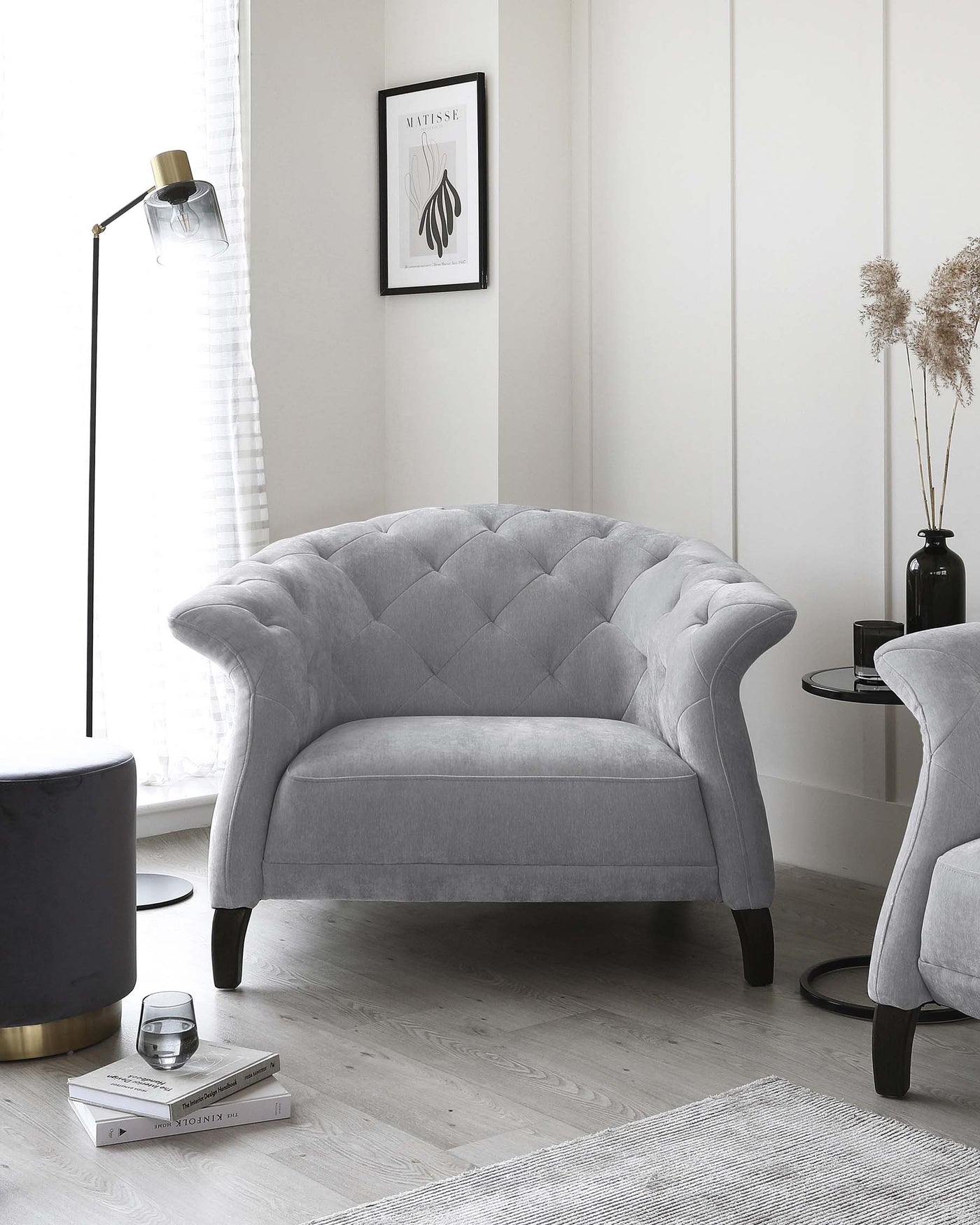 Elegant grey tufted loveseat with a curved backrest and rolled arms, featuring plush upholstery and dark wooden legs, placed on a light wooden floor, accompanied by a contemporary black and gold side table.