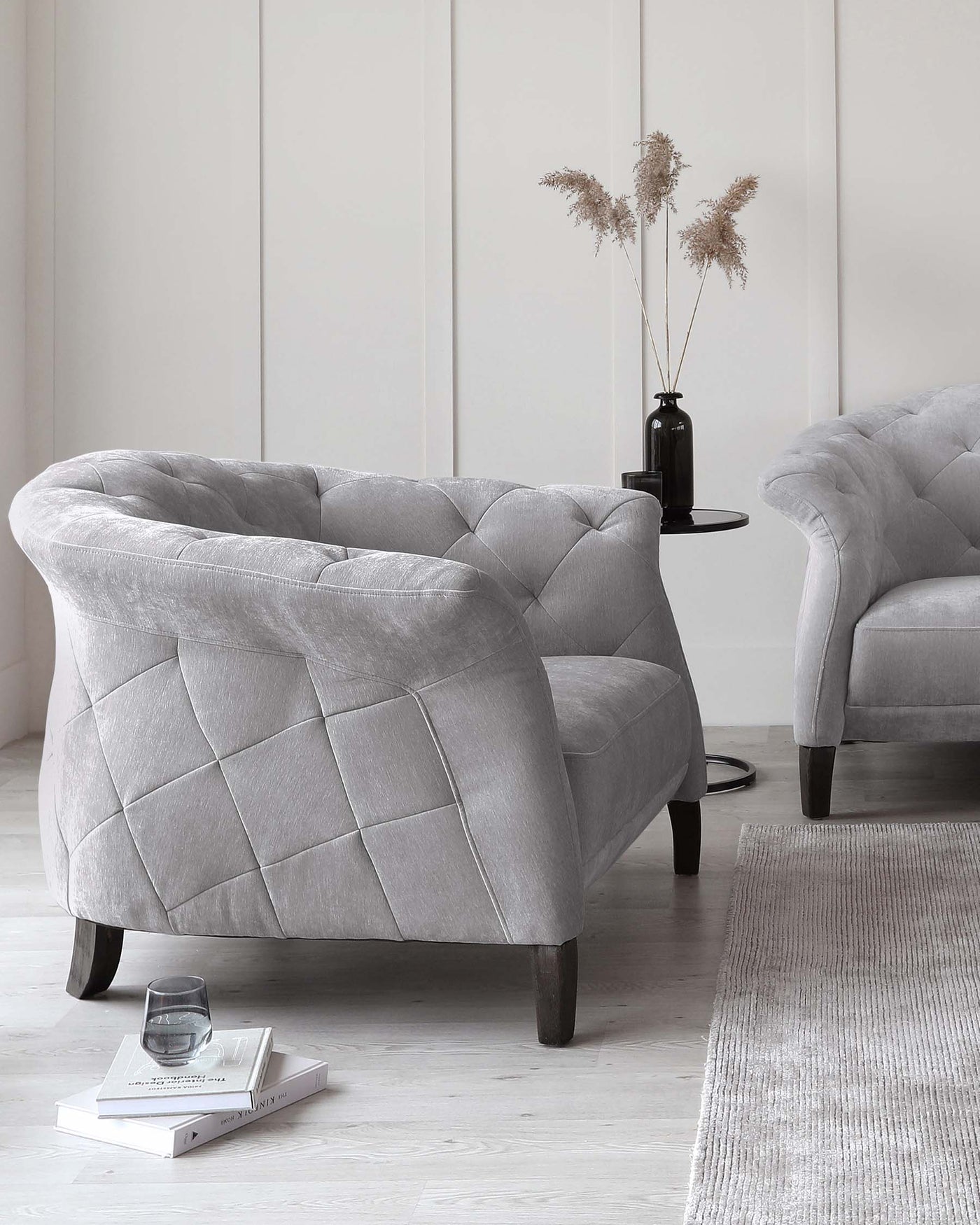 Elegant grey tufted armchairs with curved backrests and dark wooden legs, paired with a minimalistic black side table with a vase of pampas grass and positioned on a light grey textured rug.