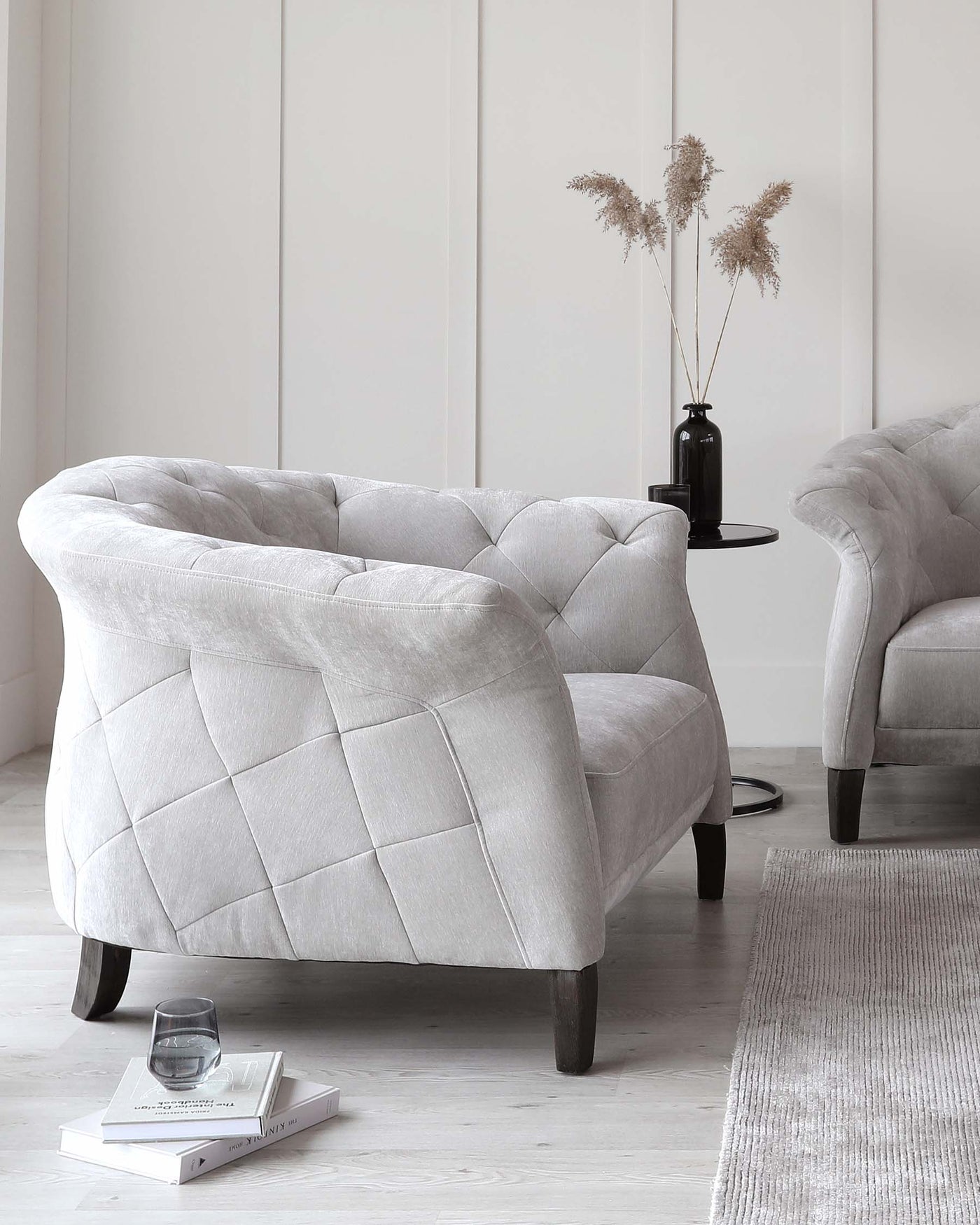 Elegant light grey tufted armchair with a voluptuous design and dark wooden legs, complemented by a neutral-toned area rug, a round side table with a vase of dried pampas grass, and a simplistic book arrangement on the floor.