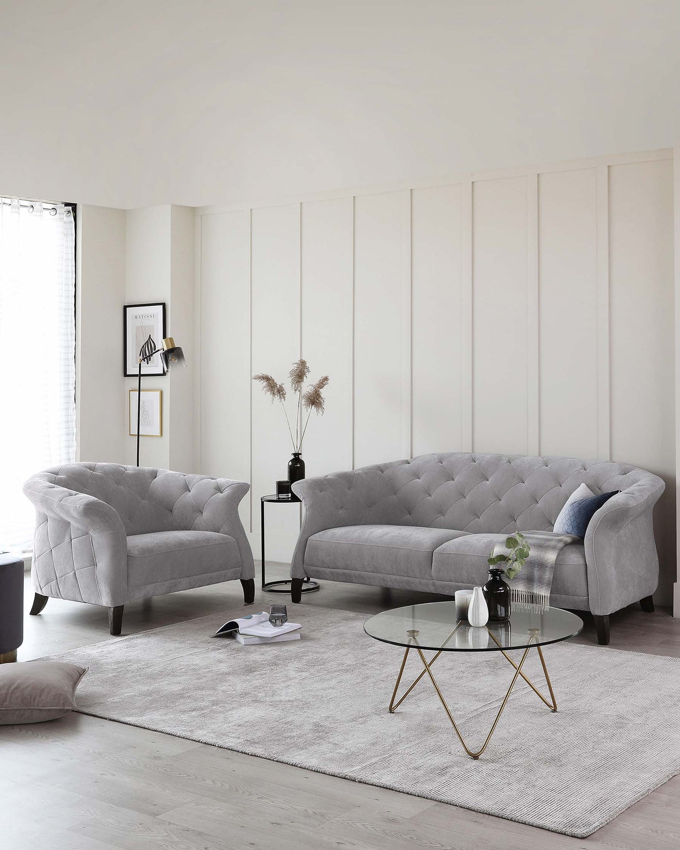 Elegant living room featuring a grey tufted sofa with a curved back, complemented by a matching tufted accent armchair. A round coffee table with a glass top and unique gold-coloured metal base is centred on a textured light grey area rug.