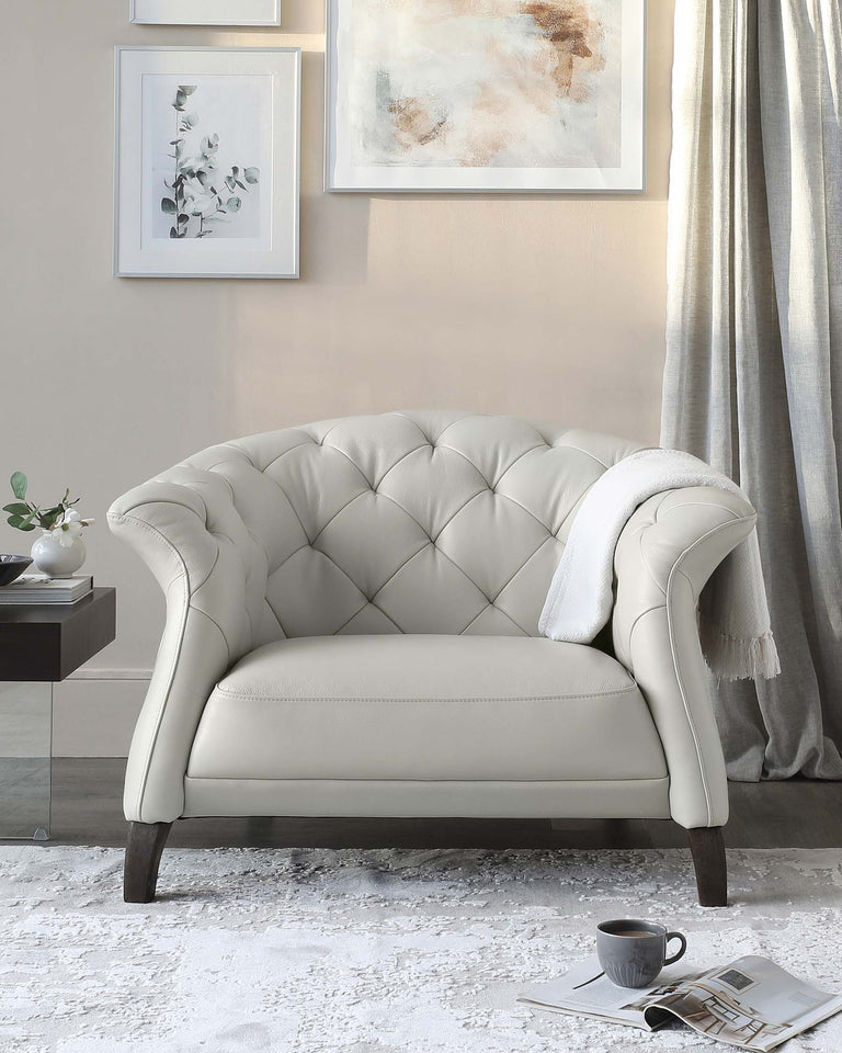 Elegant ivory tufted armchair with curved backrest and dark wooden legs, accompanied by a soft white throw blanket draped over its side. A contemporary styling with a neutral colour palette is presented in a room with soft grey curtains, abstract wall art, and a small side table with a vase. The plush white area rug beneath the chair adds a cosy texture, and a cup of coffee with a magazine beside the chair suggests a relaxed, luxurious lifestyle.