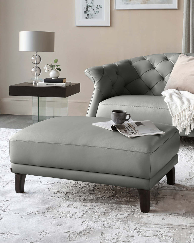 Elegant contemporary living room furniture set including a tufted grey leather loveseat, a grey rectangular leather ottoman with dark wooden legs, a glass side table with a dark wooden base, and a modern silver table lamp. The arrangement is tastefully accessorized with a pale pink throw, a small plant, books, and a cup and saucer, all atop a textured off-white area rug.