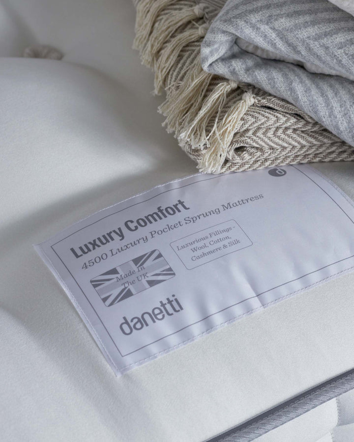 A close-up view of a white luxury pocket sprung mattress, with a corner label displaying the brand and product details. A grey textile, possibly a throw or a blanket, with a beige fringe edge, is draped over the top.