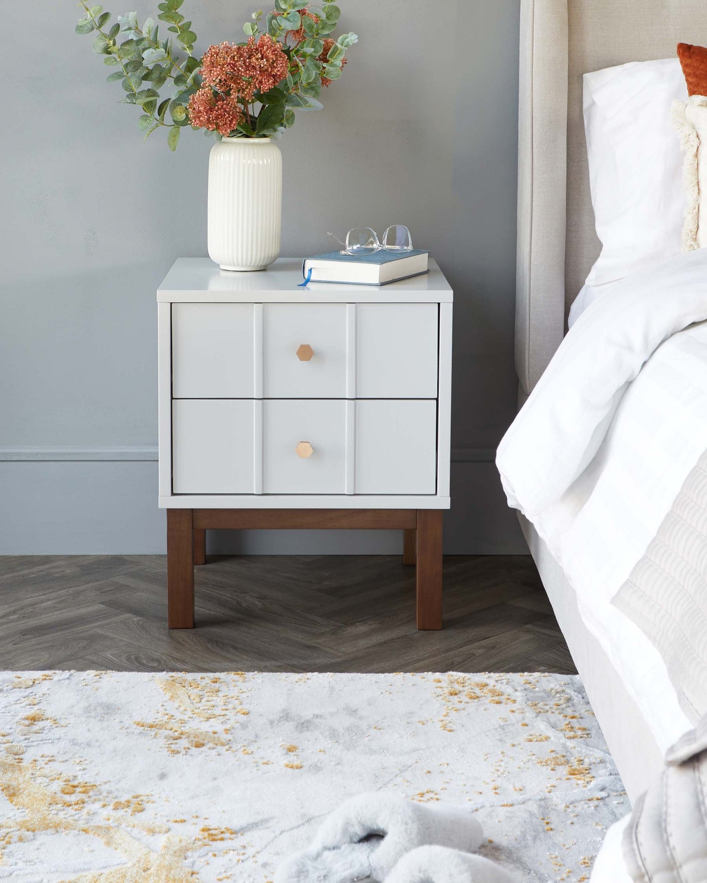 Modern minimalist white bedside table with a wooden leg base, featuring two drawers with round wooden knobs. Positioned next to a bed with white linen, the table holds a vase of flowers, a pair of glasses, and a book. A plush grey and yellow patterned rug is partially visible on the floor.