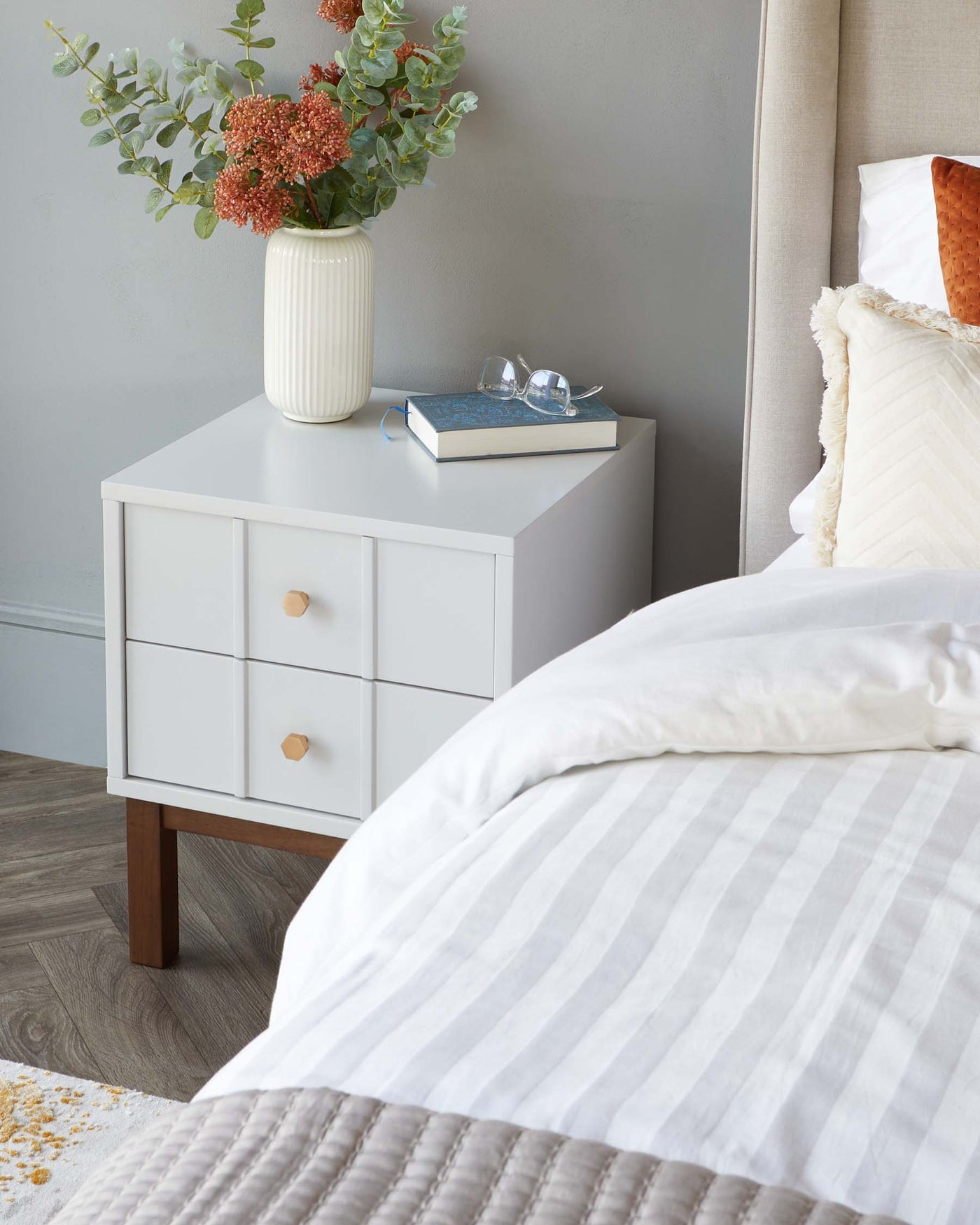 Modern white bedside table with three drawers featuring round wooden knobs, set against a grey wall, next to a bed with striped bedding.