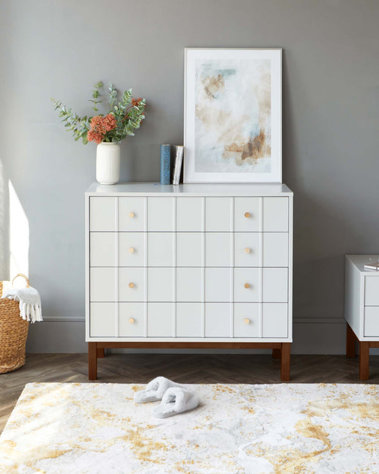 Modern white dresser with square drawers and round wooden knobs, featuring tapered wooden legs. A matching white nightstand with two drawers is visible to the right. A textured area rug with cream and beige colours is partially visible in the foreground.