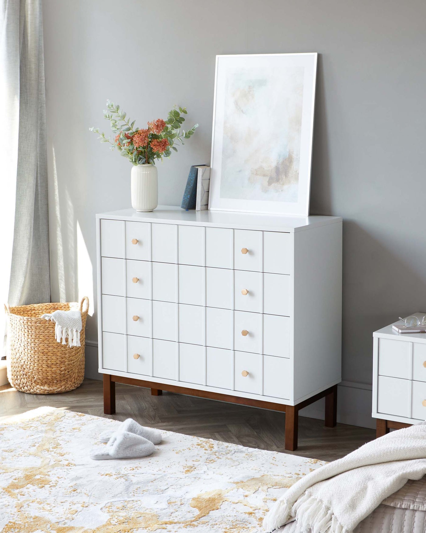 A modern white dresser with multiple drawers and round wooden knobs, standing on a minimalistic wooden base frame. To the right is a smaller, matching white bedside table with three drawers and similar wooden knobs. Both pieces are part of a contemporary bedroom furniture set.
