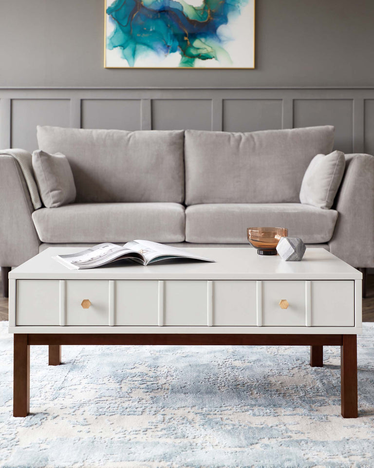 A contemporary living room setup featuring a light grey upholstered three-seater sofa with plush cushions and a sleek, rectangular off-white coffee table with drawers and wooden legs. The table is styled with minimalistic decor, including an open magazine, a small bowl, and a geometric object. The furniture rests on a soft, textured area rug with shades of blue and cream, complementing the muted tones of the room. The wall behind the sofa is adorned with an abstract painting with swirls of blue and green.