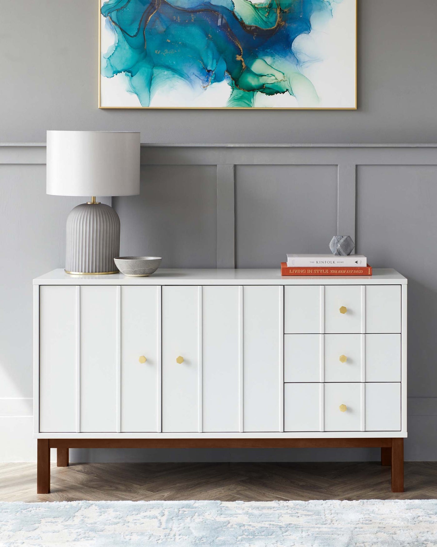 Modern white sideboard with clean lines, featuring gold handles, a mix of cabinet doors and drawers, and a contrasting dark wooden base. A contemporary lamp and decorative items are placed on top.
