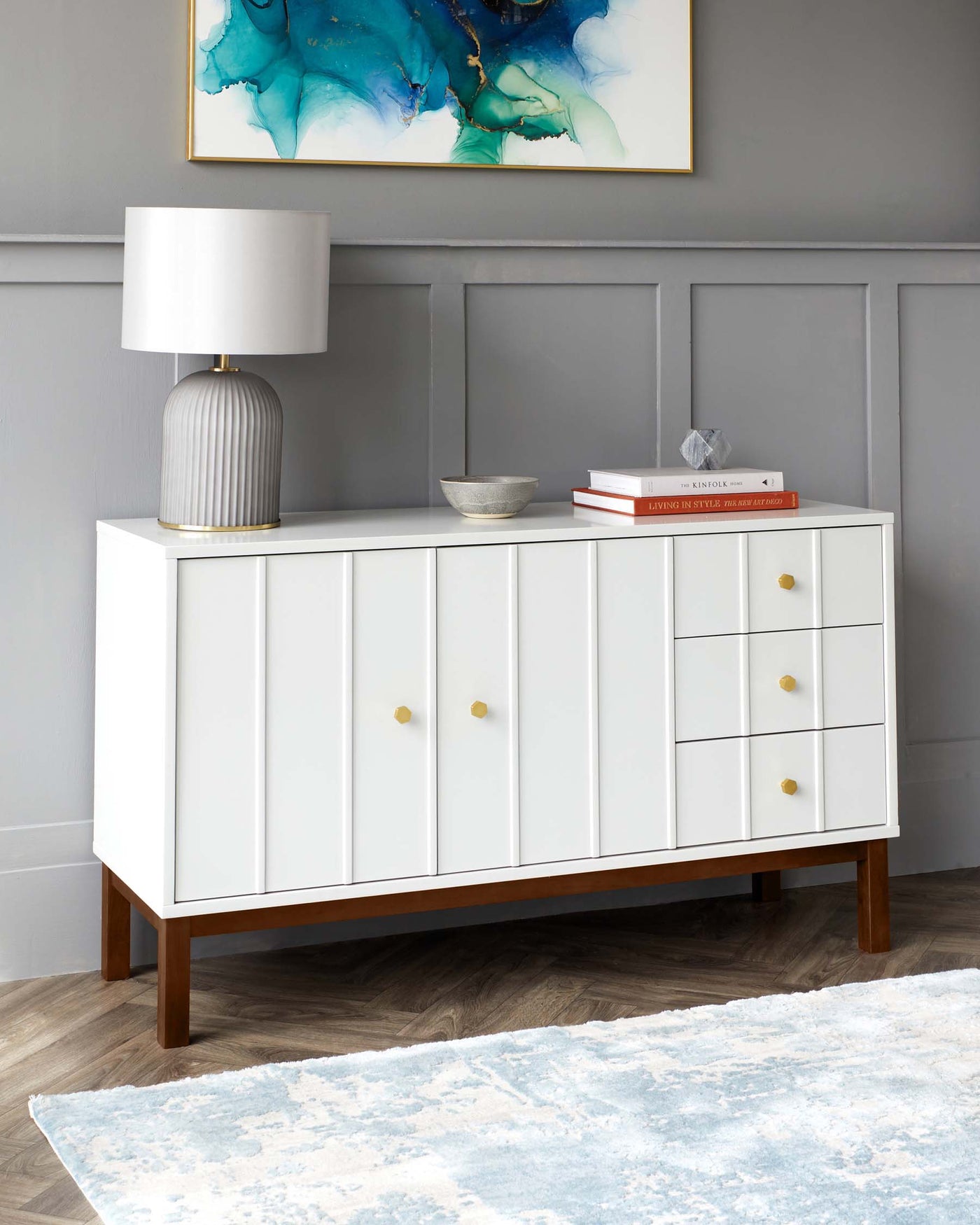 Contemporary white sideboard with golden knobs, featuring two cabinet doors and three drawers, set on tapered wooden legs. Accessorized with a grey textured lamp, a decorative silver bowl, and a stack of books atop it. The sideboard is against a wall with a modern abstract painting in blue tones, above a pale blue and white patterned area rug on a wooden floor.