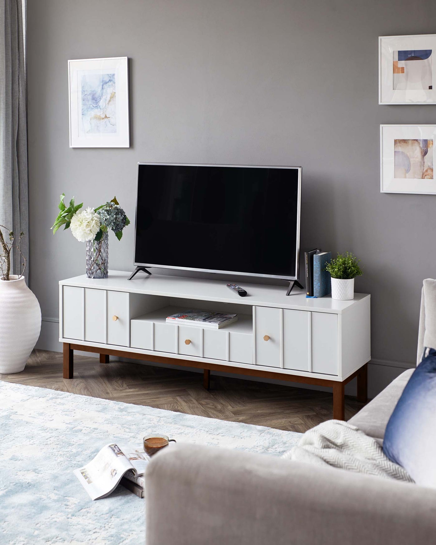 A modern white television stand with a rectangular shape and clean lines, featuring a series of drawers and open shelves. The stand has a contrasting dark wood base with tapered legs, complementing the minimalist design. A flat-screen TV is centred on top, with decorative items such as books and plants placed around it to create a homey atmosphere.