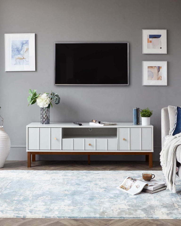 Modern minimalist white media console with gold handles, standing on splayed wooden legs, in a living room setting with a grey wall backdrop.