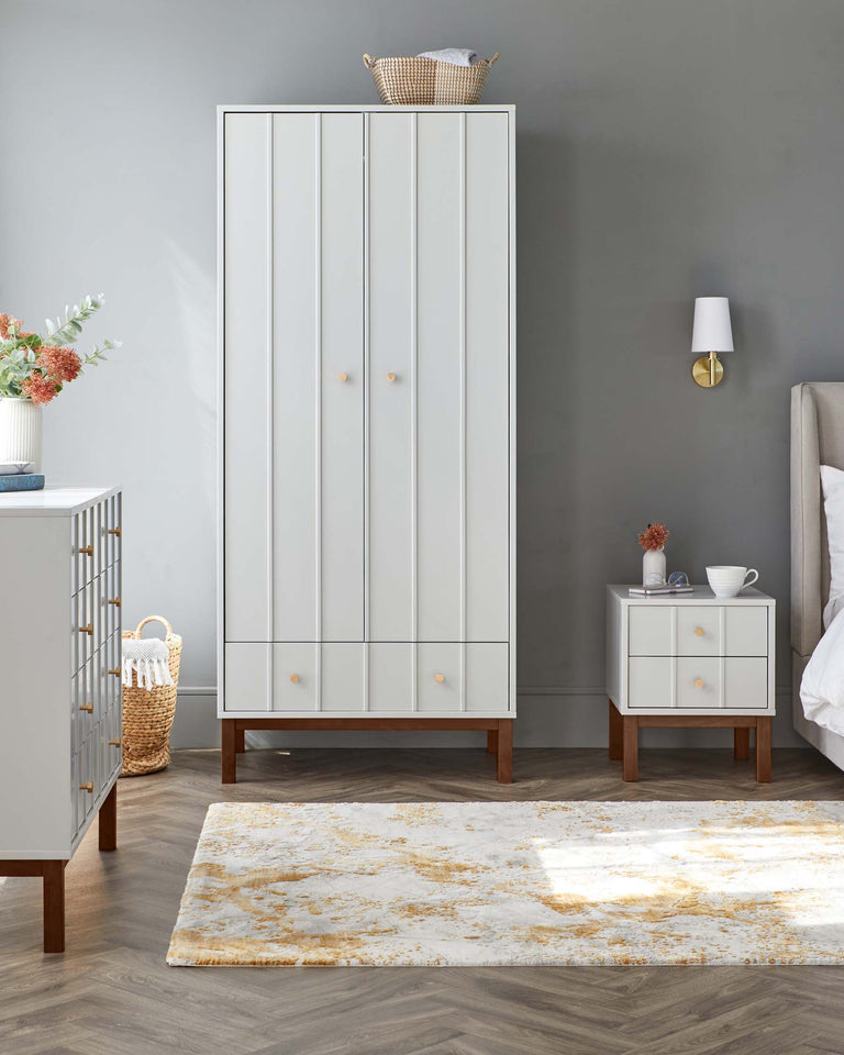A modern bedroom setting featuring a tall white wardrobe with clean lines and wooden tapered legs, complemented by a matching white two-drawer nightstand with the same wooden leg style. A decorative area rug with a white and gold abstract pattern is partially shown in the foreground.
