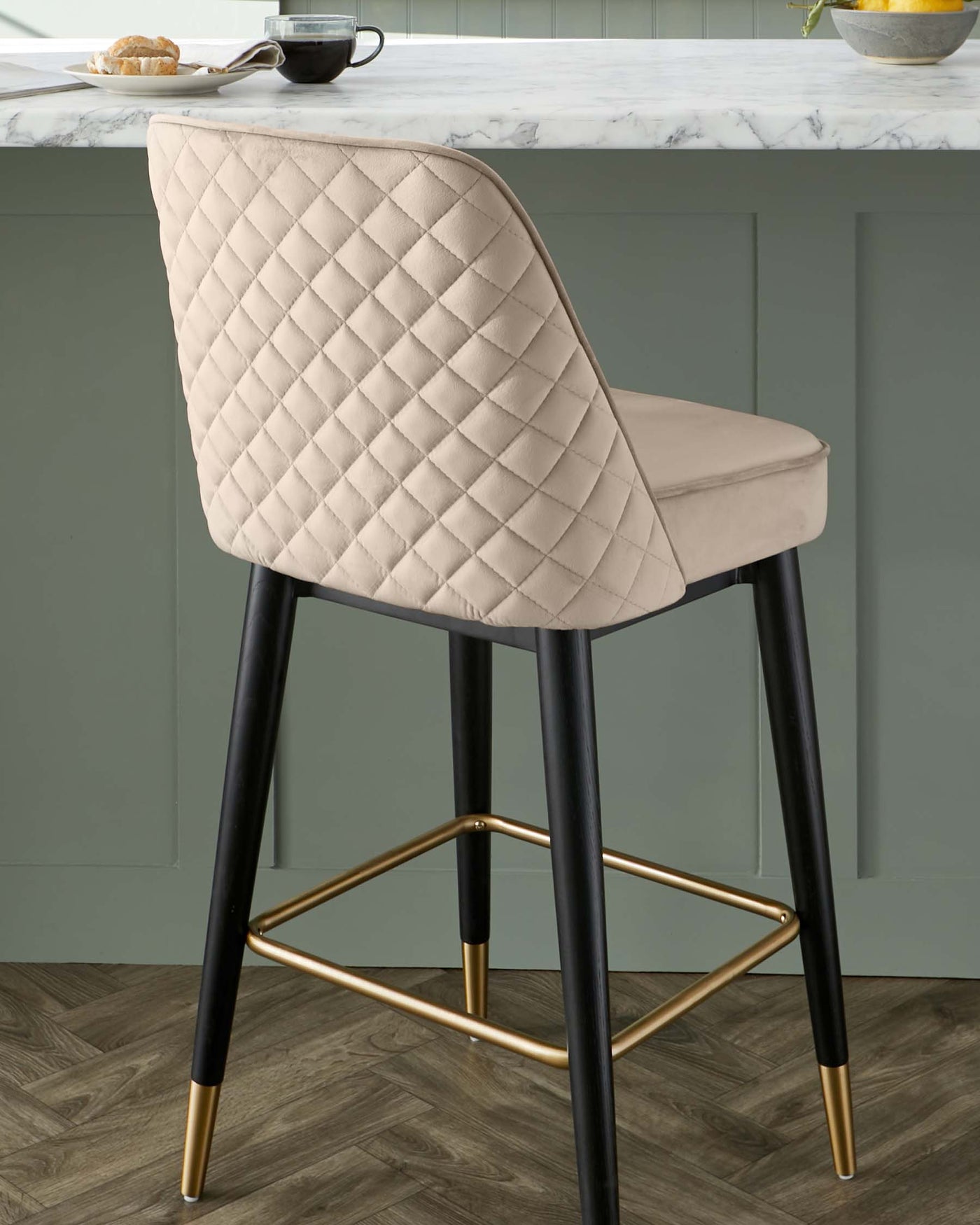 Elegant contemporary bar stool featuring a plush, beige, diamond-tufted upholstered backrest and seat, with sleek black legs accented with gold tips and a matching gold footrest.