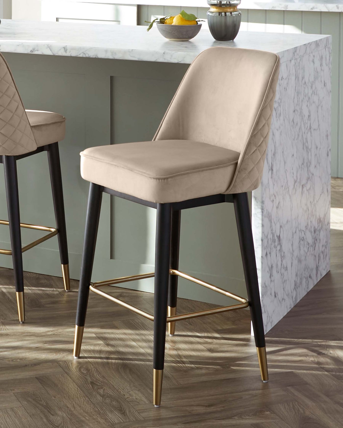 Elegant modern bar stool with a beige upholstered seat and backrest featuring diamond tufting, contrasting with sleek black legs tipped with gold accents. The stool sits beside a marble kitchen island on a wooden floor, suggesting a luxurious and contemporary aesthetic.
