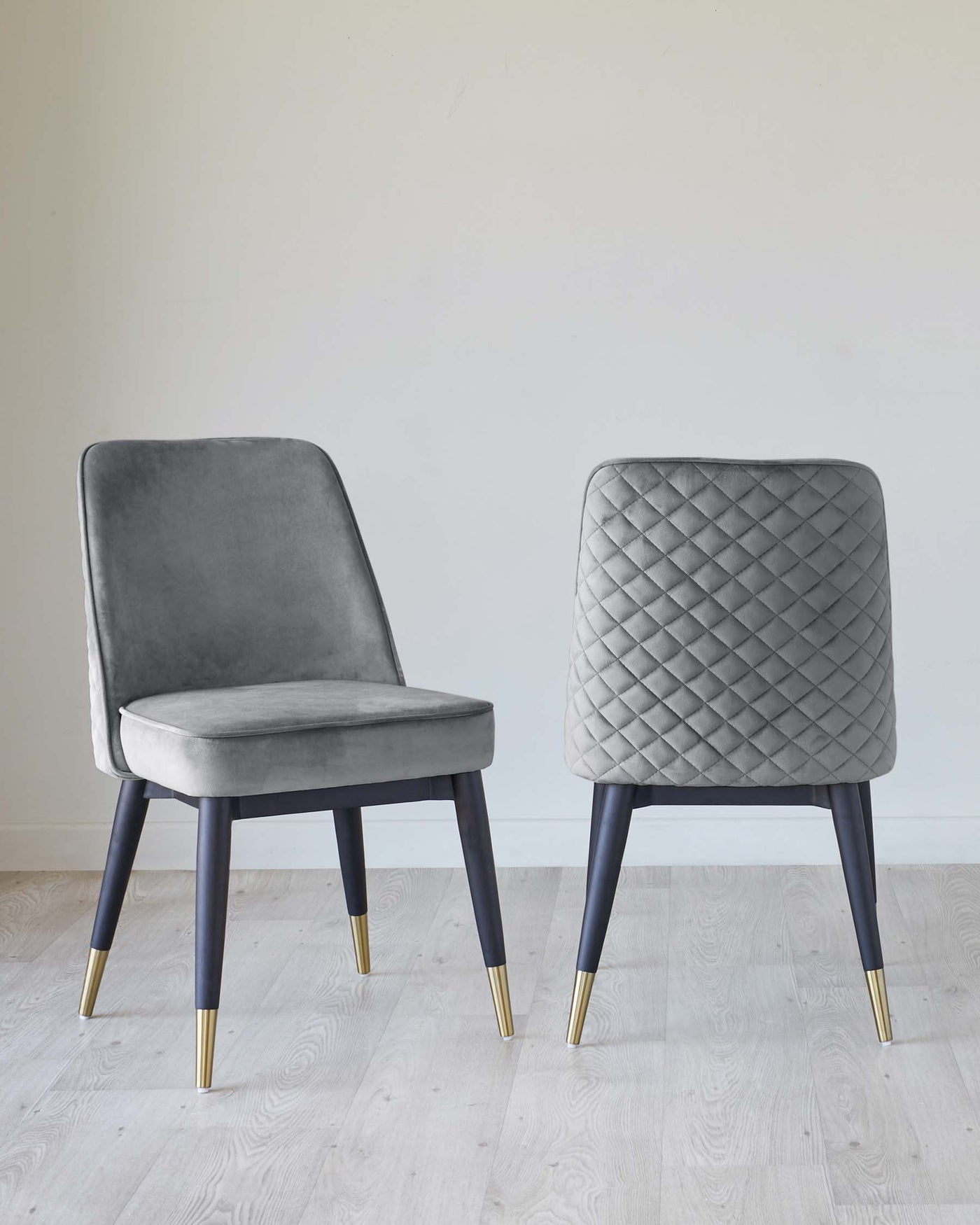 Two modern dining chairs with elegant velvet upholstery in grey, featuring one with a smooth finish and the other with a diamond tufted backrest. Both chairs have dark wooden legs accented with gold tips, set against a light wooden floor and a clean white wall backdrop.