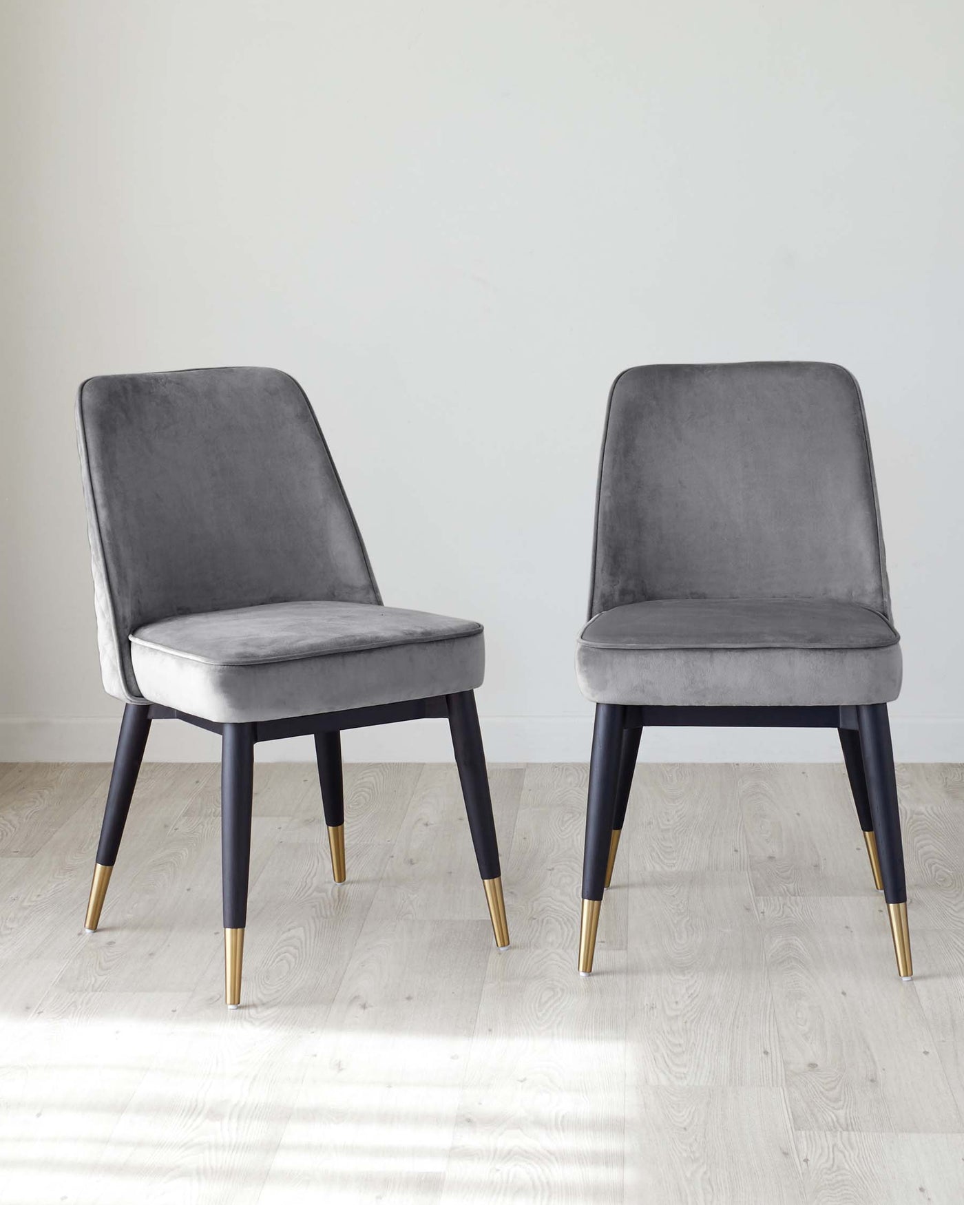 Two modern dining chairs with elegant grey velvet upholstery and dark wood legs tipped with gold accents, showcased on a light hardwood floor against a neutral background.