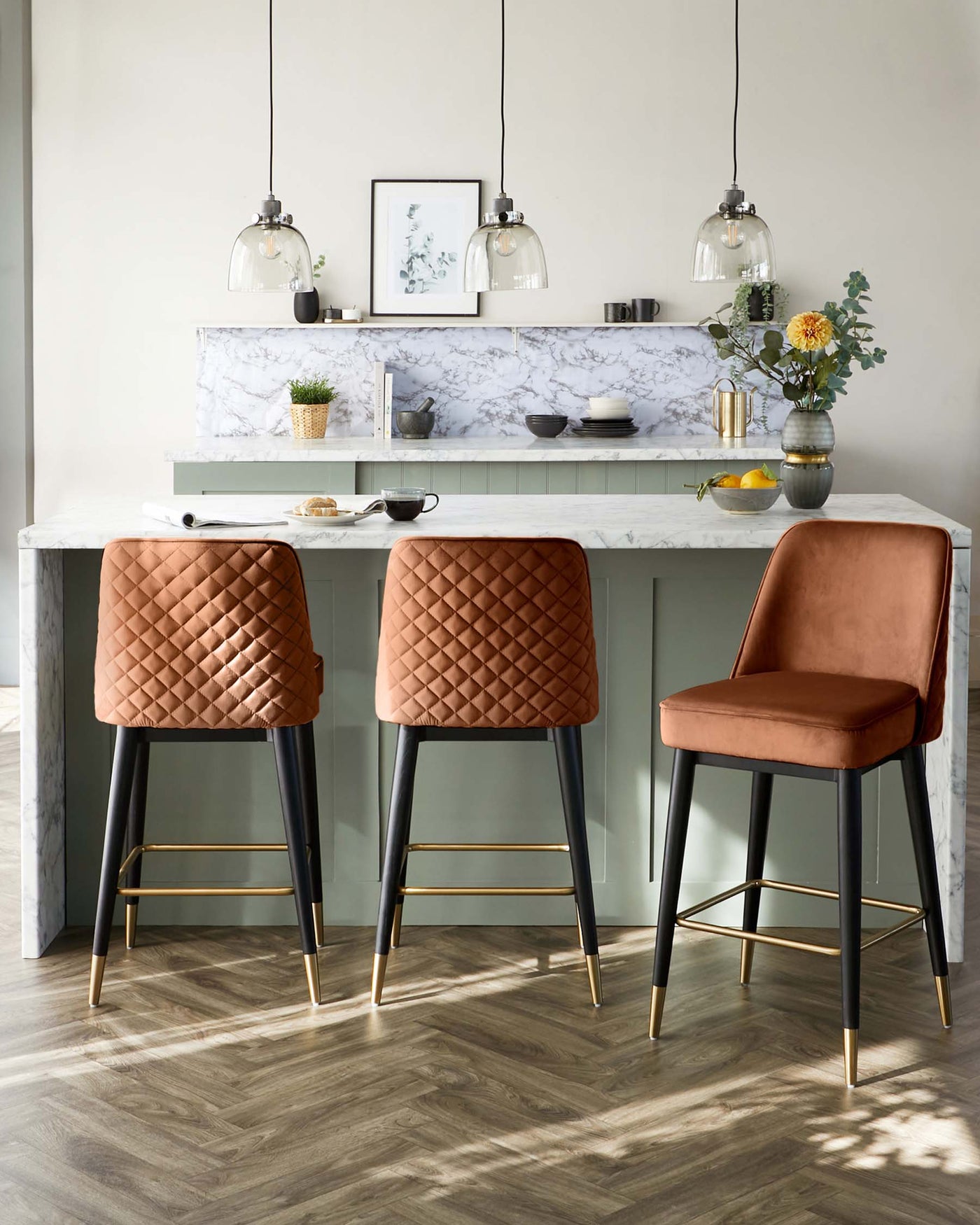 Modern kitchen bar setting featuring three stylish bar stools with quilted leather upholstery in shades of caramel and a single stool in burnt orange, all with slender black legs accented with gold tips. A marble-topped kitchen island with a pale green base serves as a breakfast bar, complemented by minimalist pendant lights overhead.