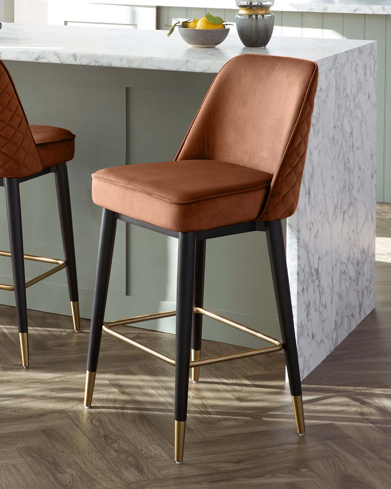Elegant modern bar stools with a caramel-coloured, quilted leather upholstery on the seat and back, supported by a sleek black frame with brass-finished footrests and leg tips, displayed in a contemporary kitchen setup with marble countertops.