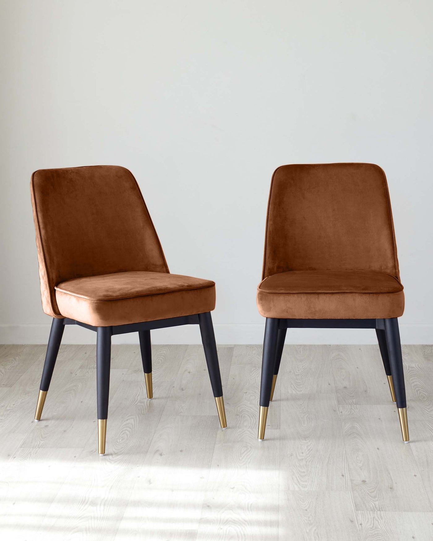 Two modern dining chairs with velvety caramel upholstery and sleek black legs tipped with gold accents, set against a simple grey background on a light wooden floor.