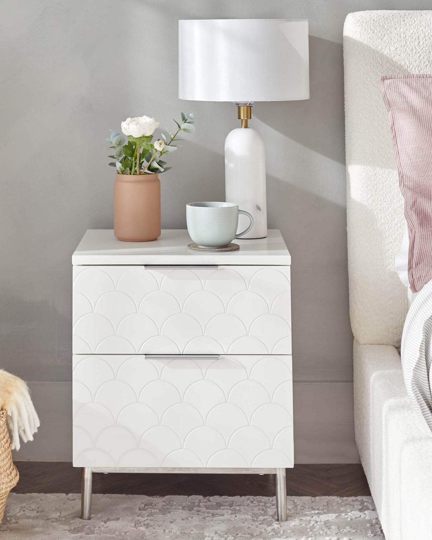 A modern white two-drawer nightstand with a scalloped pattern on the drawer fronts, resting on sleek metal legs. The nightstand is accessorised with a white cylindrical lamp with a large white shade, a terracotta vase with white flowers, and a pale blue mug on a coaster. The piece is positioned next to a light-coloured upholstered bed with a textured throw blanket, set against a light grey wall on a plush off-white carpet.