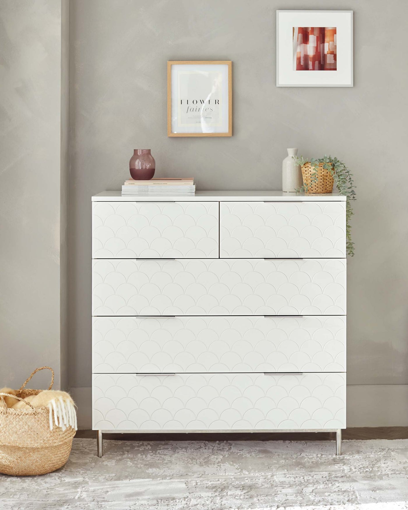Modern white six-drawer dresser with a scalloped pattern facade and minimalistic metal handles, set against a grey wall with decorative items on top and a woven basket beside it.