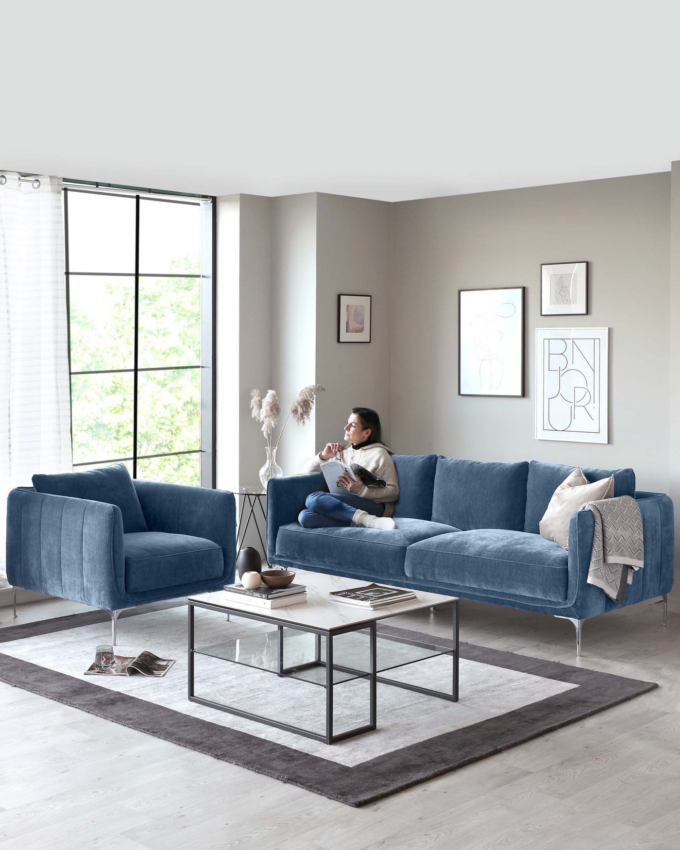Modern living room furniture in plush blue fabric consisting of a three-seater sofa with clean lines, cylindrical cushions, and metal legs, accompanied by a matching single-seater armchair. A square metal-framed coffee table with a glass top sits on a grey area rug, and a side table with a round glass top and a black tripod base is present.