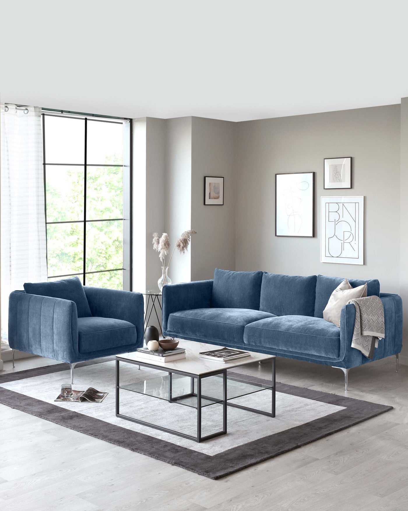 Modern living room setup featuring a plush blue sectional sofa with squared cushions, complemented by a matching blue loveseat. A rectangular glass-top coffee table with a minimalistic metal frame is centred on a grey area rug. The space is accented with decorative throw pillows, a soft blanket, and elegant wall art, creating a cosy and stylish atmosphere.