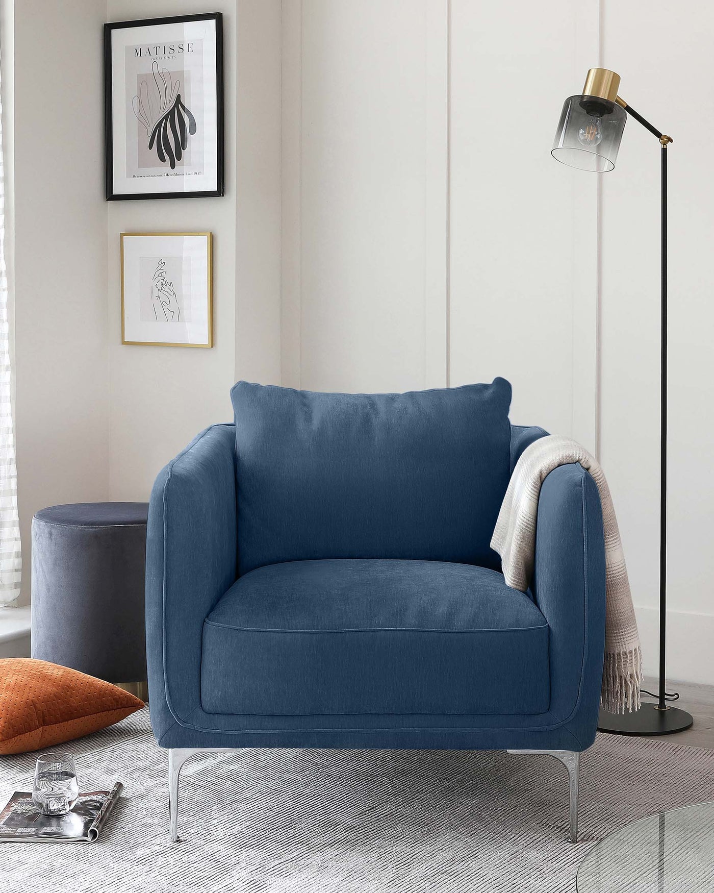 A modern navy blue fabric armchair with clean lines and metal legs, paired with a matching round navy blue ottoman, complemented by a sleek black floor lamp with a transparent shade and brass accents.
