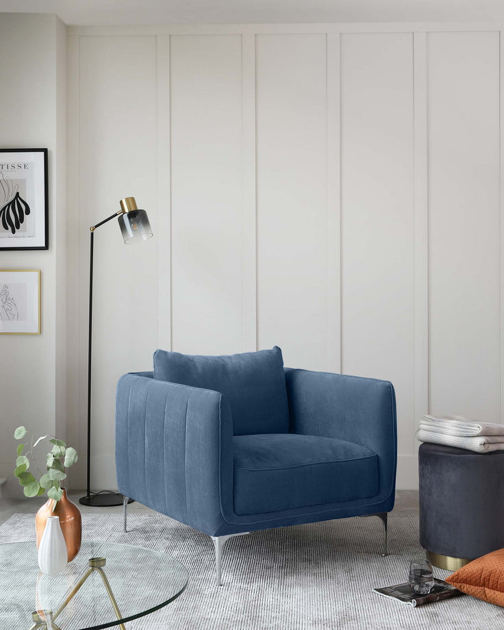 A contemporary blue velvet loveseat with vertical channel tufting and sleek metallic legs; a round glass-top side table with a gold-finished base; and a round, dark blue upholstered ottoman with a hidden storage feature and a brass base.