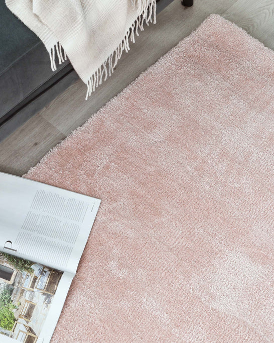 Plush, pale pink area rug positioned on a wooden floor, partly visible beneath an open magazine, with the edge of a light-coloured throw blanket hanging over the side, hinting at cosy, modern living space furnishings.