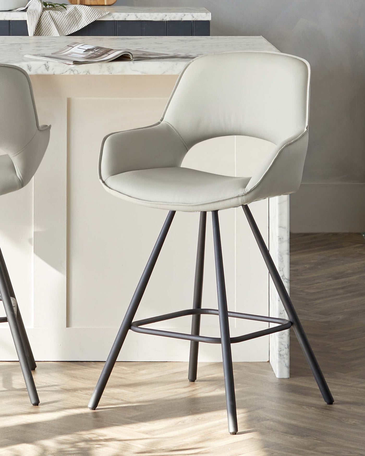Modern light grey upholstered bar stool with a sweeping curved backrest and a comfortable, rounded seat pad, mounted on a sleek black metal frame with four slender legs and a circular footrest. The stool is positioned near a kitchen island with a white marble countertop.