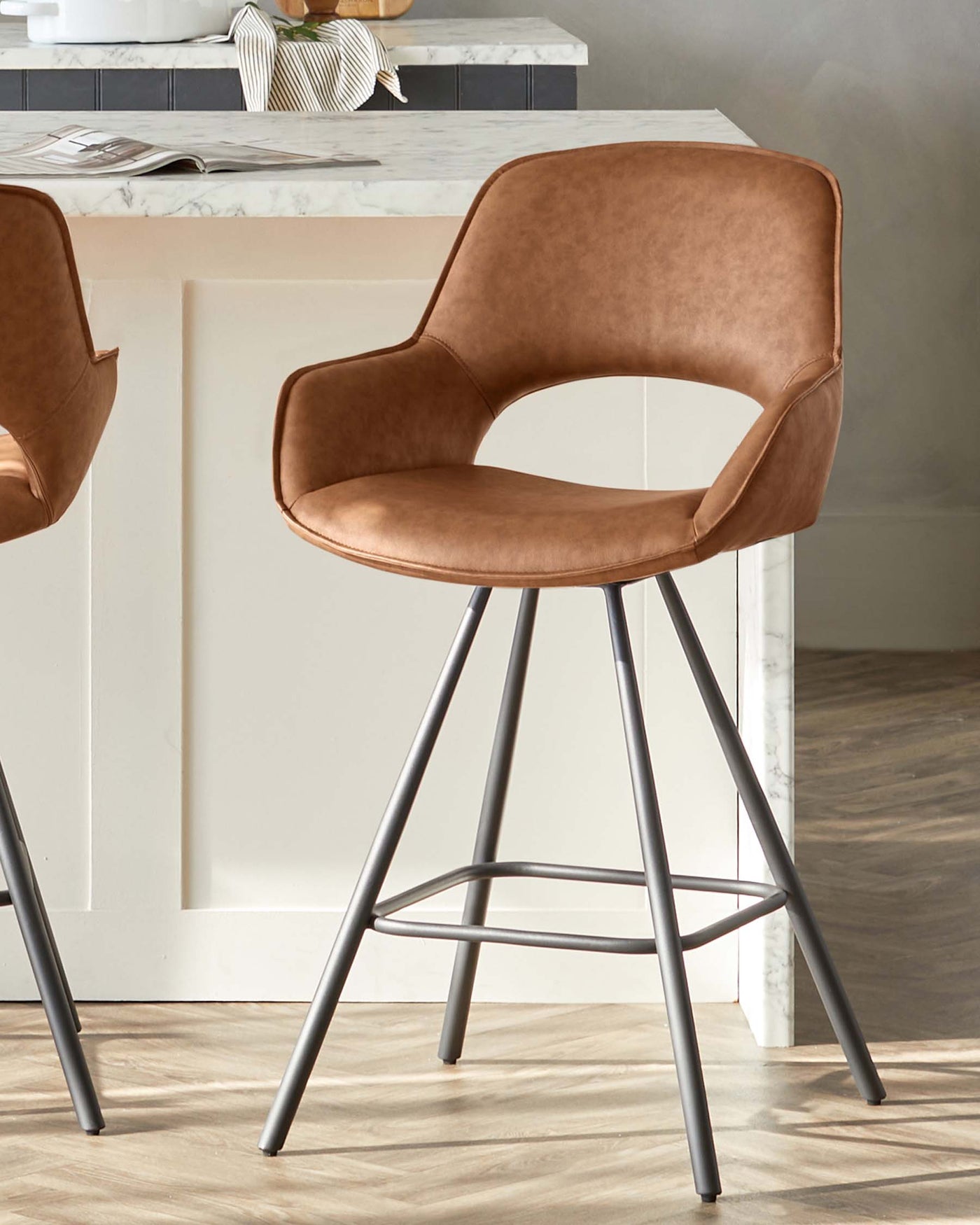 Elegant modern bar stool with a smooth caramel brown faux leather upholstery and curved backrest, featuring a minimalistic black metal frame with four legs and footrest for added comfort.