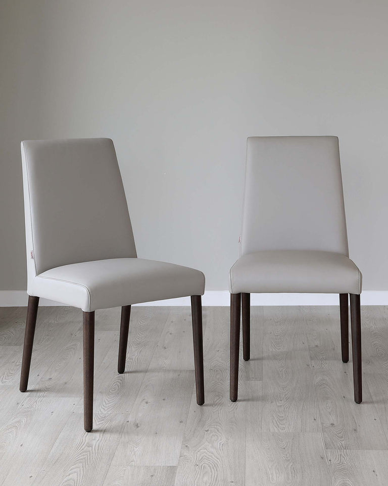 Two modern minimalist dining chairs with light grey upholstered seats and backs, featuring dark wooden legs. The chairs are positioned on a light wooden floor against a plain light grey wall, highlighting their sleek and contemporary design.
